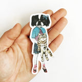 Hand holding dog sticker. Cute dressed up dog in sweater, skirt, and shoes, holding a fluffy kitty