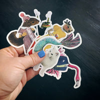 Holding various animal stickers, mermaid cat sticker, illustrated squirrel sticker, otter vinyl sticker, flamingo dude sticker, alligator retro sticker