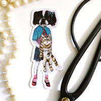 Original illustration sticker of dog and cat friends. Dressed up dog is holding a cat. Pictured with pearls and scissors.