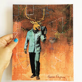 Hand holding an 8x10" art print. Mixed media illustration of vintage dressed buck on a brown background. Buck is wearing a blue suit. Vintage Inspired Wall Decor - Vintage Bar Decor - Retro Buck Art Print by Pergamo Paper Goods