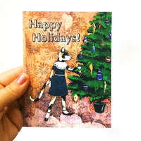 Hand holding holiday card featuring a cat putting an ornament on a Christmas tree